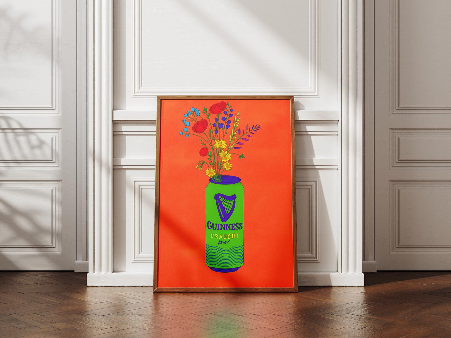 Framed artwork of a green Guinness bottle with flowers against an orange background, displayed in a room with white walls and wooden floor.