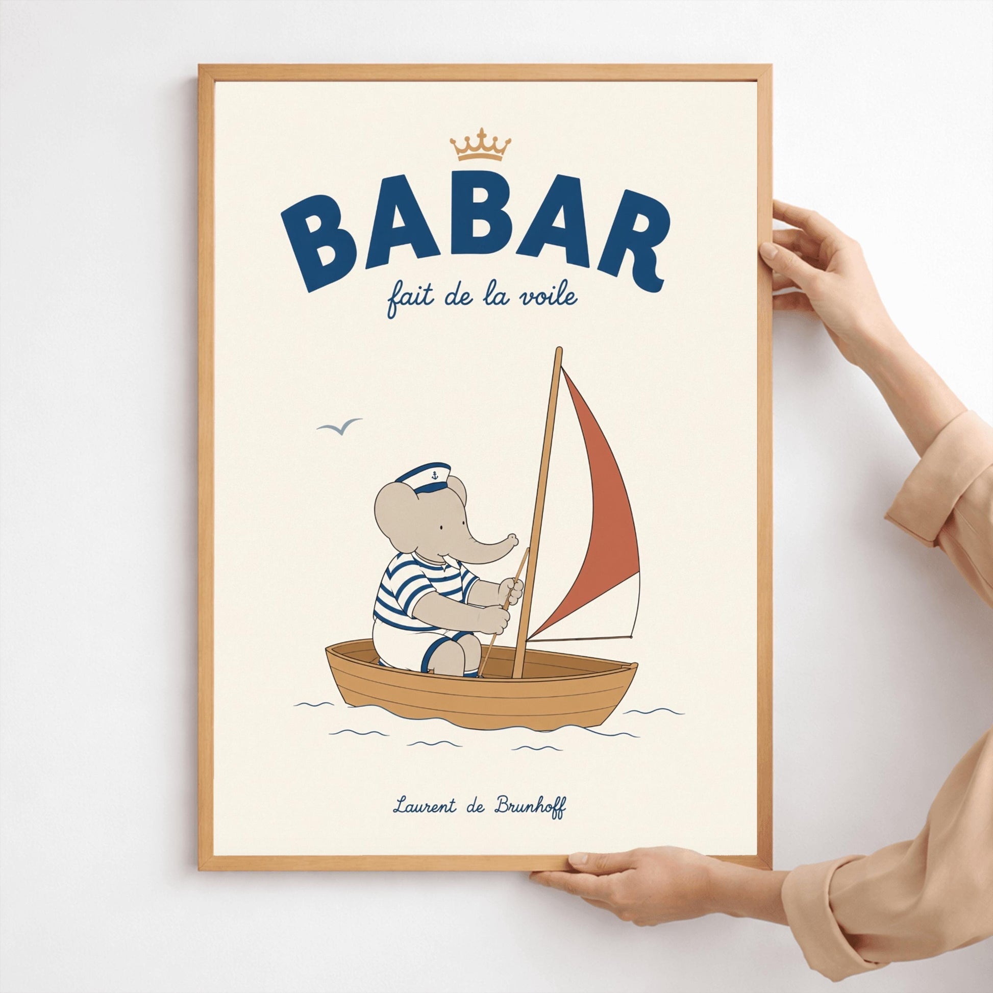 A vertical storybook-style art print featuring Babar the Elephant sailing a small boat. Babar wears a blue and white striped shirt and a white sailor's hat with an anchor. He is sitting in a brown wooden boat with a red and white sail. A single seagull flies in the creamy off-white sky. The top reads "BABAR fait de la voile" with a small gold crown.