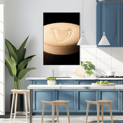 A vertical macro photograph of the thick, creamy foam head on a pint of Guinness. The iconic Guinness harp is embossed into the center of the velvety bubbles. The image features warm cream tones against a dark, shadowy background.