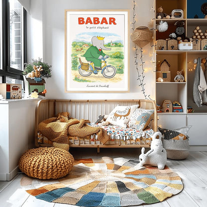 A vertical children's illustration poster titled "BABAR le petit éléphant" at the top in red. King Babar, in a green suit and crown, is riding a yellow bicycle along a dirt path. A brown leather bag on the back of the bike holds a baguette. The background shows a green meadow with a cow and calf, small bushes with flowers, and a distant house and church under a soft sky. The name "Laurent de Brunhoff" is at the bottom.