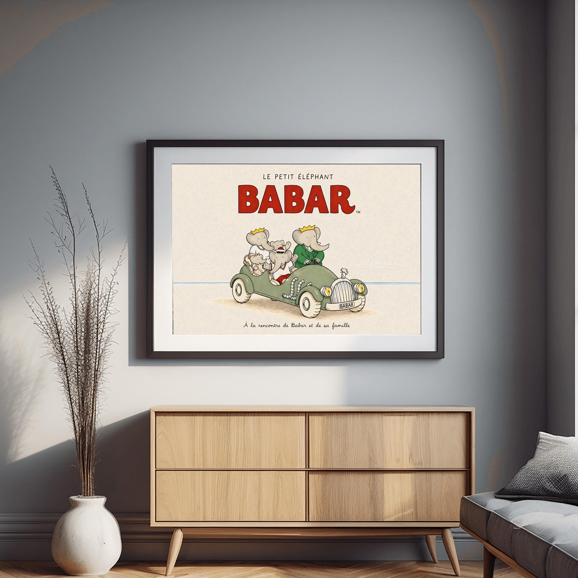 A horizontal illustration featuring Babar the Elephant and his family in a vintage sage-green roadster. Babar, wearing a green suit and crown, is driving. Beside him and in the back seat are his wife Celeste and three young elephants. The car is on a light sandy beach under a large red "BABAR" title. The background is a simple, pale cream sky with a thin blue ocean line. French text at the bottom reads, "À la rencontre de Babar et de sa famille."