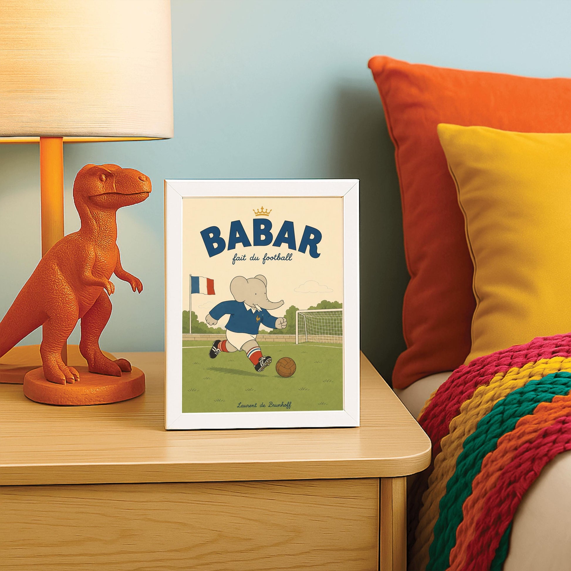 A vertical storybook-style art print featuring Babar the Elephant playing soccer on a green field. Babar wears a blue jersey with a small gold rooster logo, white shorts, red striped socks, and black cleats. He is dribbling a brown leather soccer ball toward a white goal. A French flag flies in the background. The text at the top says "BABAR fait du football" with a small gold crown.