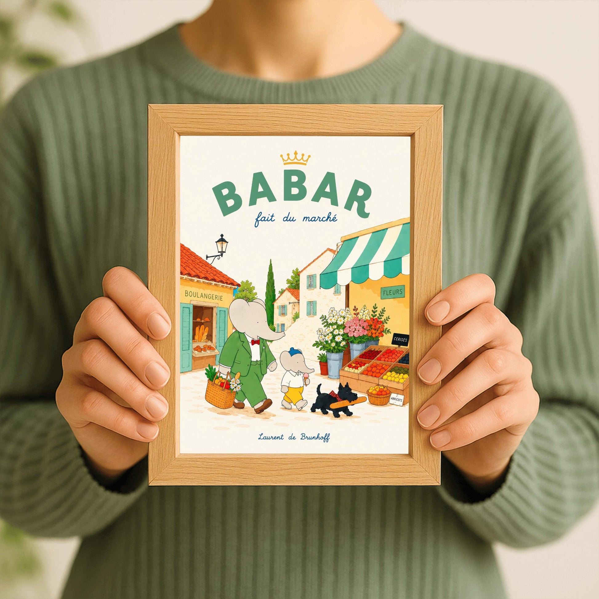 A vertical storybook-style art print of Babar the Elephant walking through a French village street. Babar, in a green suit, carries a basket of groceries. A smaller elephant walks beside him eating a pink ice cream cone, followed by a black dog carrying a baguette. The background features a boulangerie with a yellow facade, a flower stall with pink and white roses, and terracotta-roofed houses under a cream sky. The top reads "BABAR fait du marché" with a gold crown.