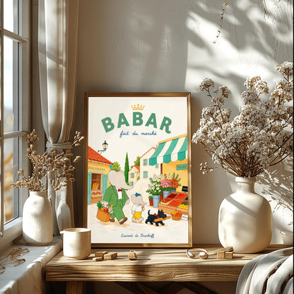 A vertical storybook-style art print of Babar the Elephant walking through a French village street. Babar, in a green suit, carries a basket of groceries. A smaller elephant walks beside him eating a pink ice cream cone, followed by a black dog carrying a baguette. The background features a boulangerie with a yellow facade, a flower stall with pink and white roses, and terracotta-roofed houses under a cream sky. The top reads "BABAR fait du marché" with a gold crown.