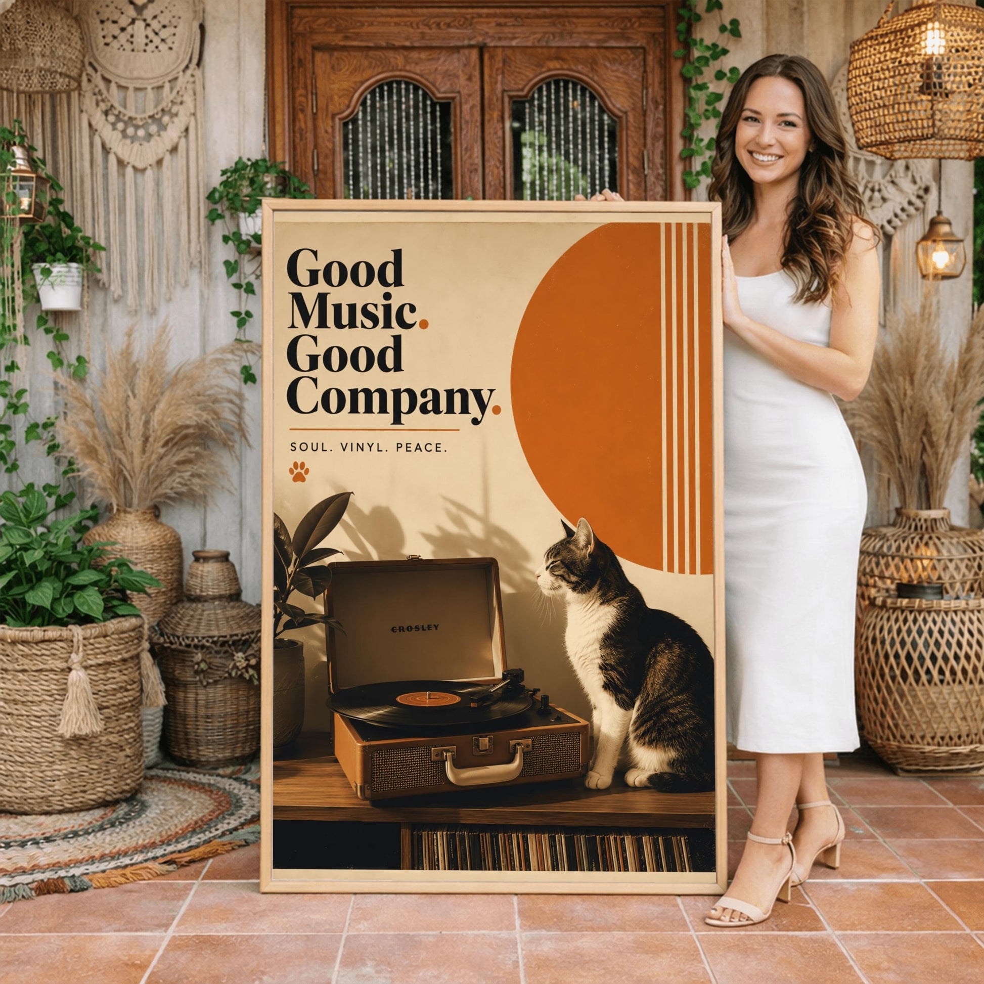 A vertical retro fine art poster titled "Good Music. Good Company." on a wide cream border. The design features a bold black serif heading on the left above the text "SOUL. VINYL. PEACE." and a small orange paw print. In the central artwork, a black and white tuxedo cat sits contentedly next to an open tan Crosley record player spinning a vinyl record. A large orange sun shape with white vertical stripes and a potted rubber plant decorate the background.