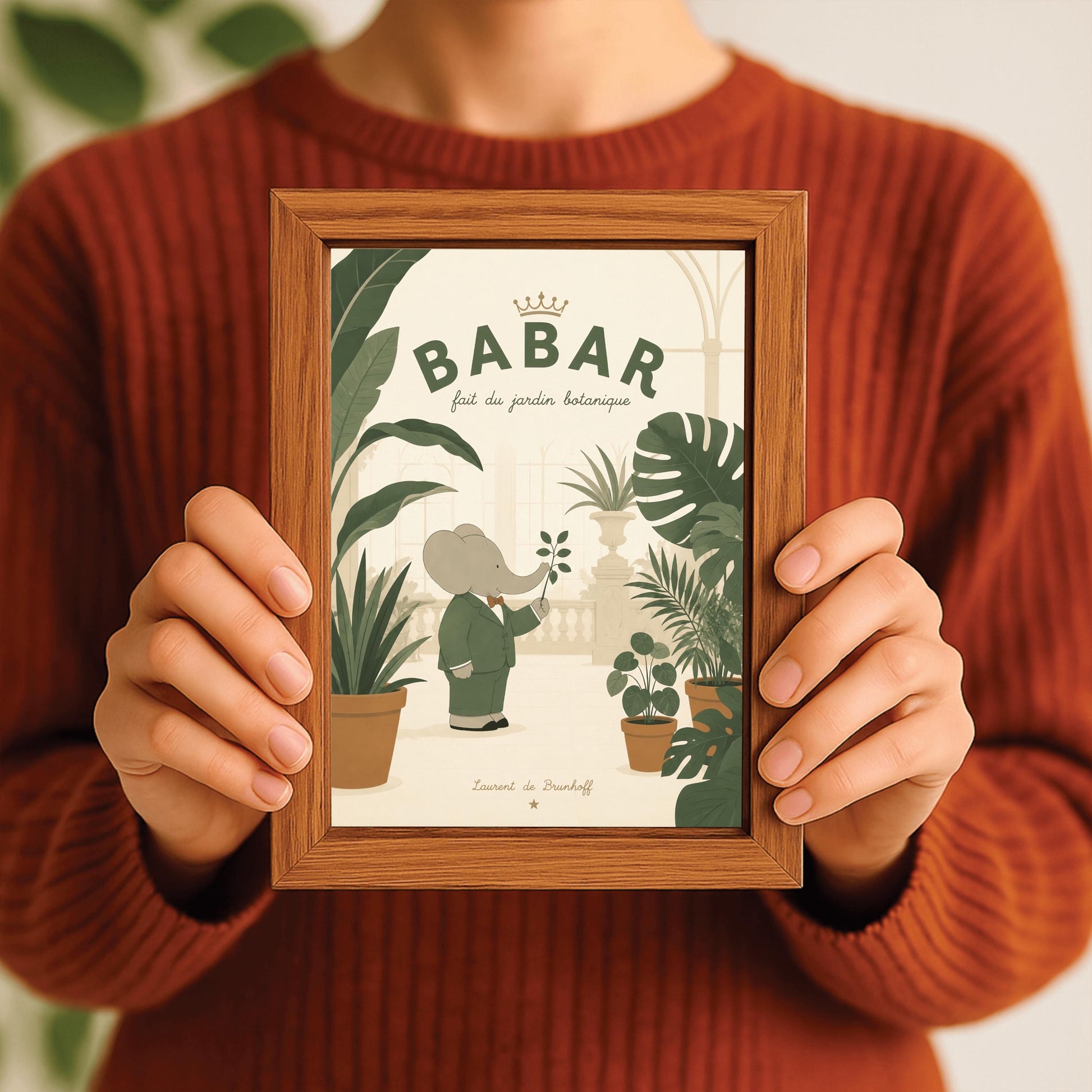 A vertical storybook-style art print of Babar the Elephant in a botanical garden. Babar, in a green suit and orange bow tie, holds a small branch while standing among various large potted tropical plants like monstera. The background shows a light cream conservatory with a glass roof and stone railings. The top reads "BABAR fait du jardin botanique" with a gold crown.