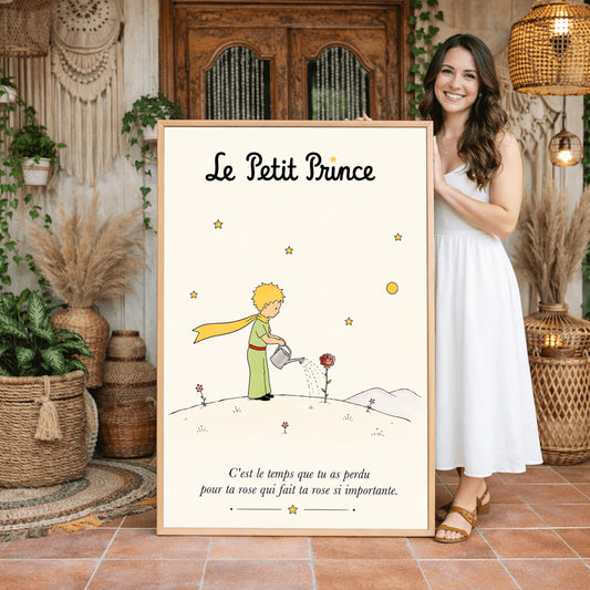 A vertical minimalist illustration poster for "Le Petit Prince" on a warm cream background. The central ink sketch shows the young boy with golden-yellow hair and a long yellow scarf leaning forward on a small hill while holding a silver watering can. Water streams down onto a single red rose blooming in front of him. Tiny red flowers grow on the ground, with soft grey hills in the distance and line-drawn stars and a yellow moon floating in the sky.