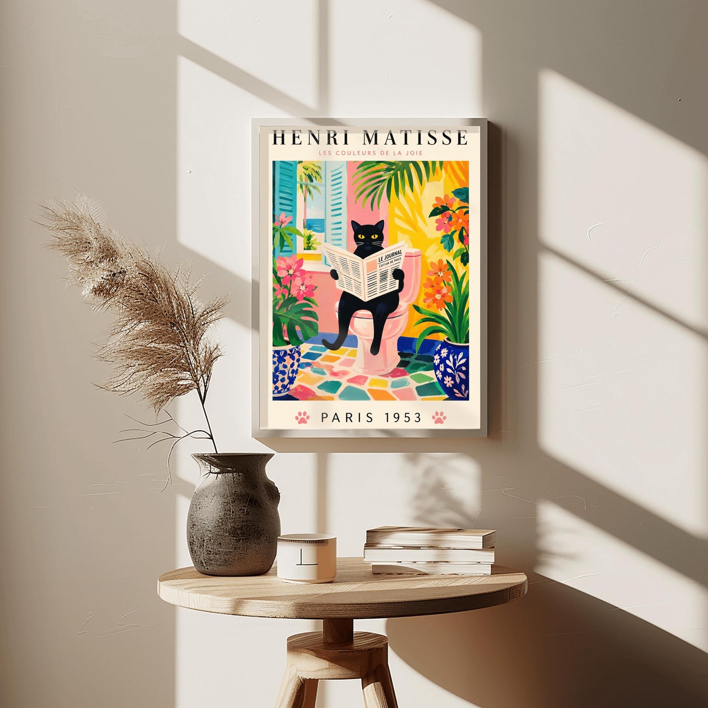 A vertical gallery poster on a cream background titled "HENRI MATISSE - LES COULEURS DE LA JOIE" in black and pink font. The central image shows a sleek black cat with bright yellow eyes sitting on a pastel pink toilet, reading a French newspaper titled "LE JOURNAL". The bathroom features an open window with blue shutters looking out at a beach, ocean, and palm trees. The room is decorated with lush green plants, potted orange flowers, and a colorful mosaic stone floor.