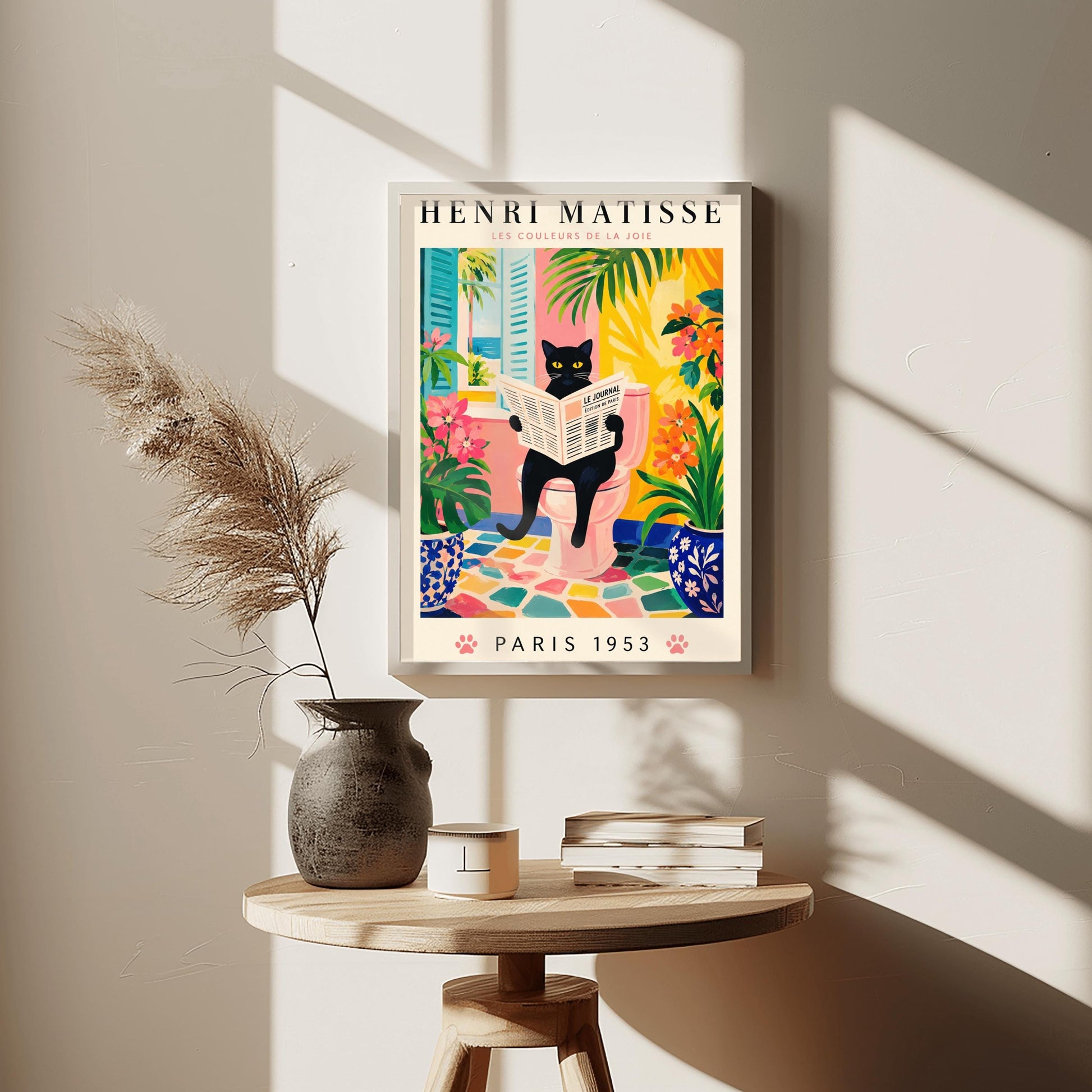 A vertical gallery poster on a cream background titled "HENRI MATISSE - LES COULEURS DE LA JOIE" in black and pink font. The central image shows a sleek black cat with bright yellow eyes sitting on a pastel pink toilet, reading a French newspaper titled "LE JOURNAL". The bathroom features an open window with blue shutters looking out at a beach, ocean, and palm trees. The room is decorated with lush green plants, potted orange flowers, and a colorful mosaic stone floor.