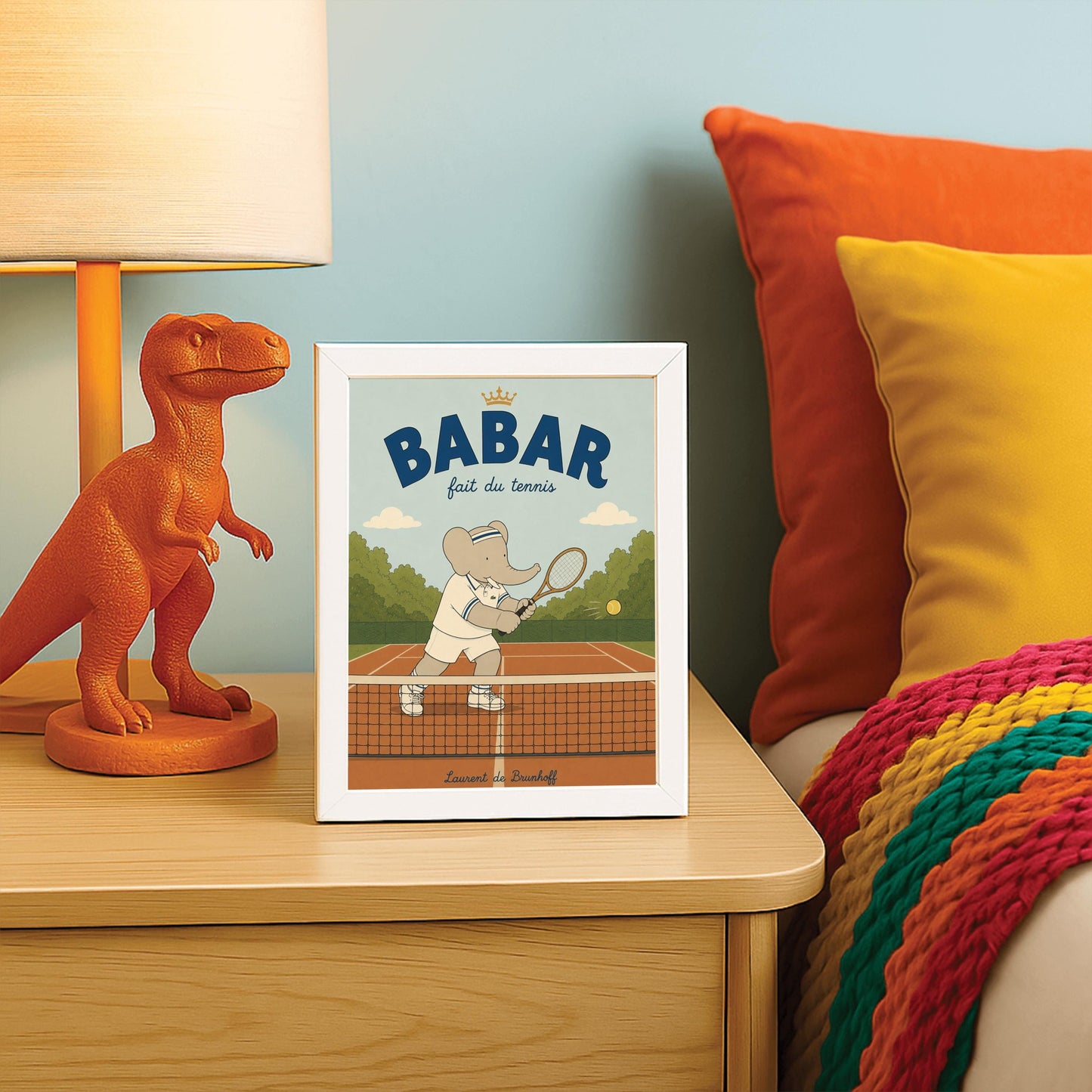 A vertical storybook-style art print featuring Babar the Elephant playing tennis. Babar is seen behind a tennis net on an orange clay court, holding a wooden racket in a backhand swing. He wears a white polo with blue stripes, white shorts, and a blue-and-white striped headband. A yellow tennis ball is in mid-air. The background has green trees and a light blue sky. The top reads "BABAR fait du tennis" with a small gold crown.