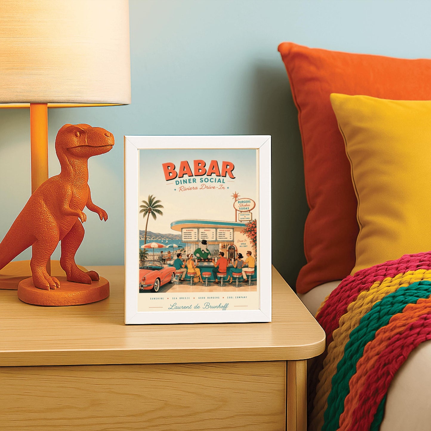 A mid-century style illustration poster titled "BABAR DINER SOCIAL - Riviera Drive-In" at the top. The scene shows an open-air retro diner bar right on the coast of the French Riviera, with a turquoise sea and white sailboats in the background. King Babar, a cartoon elephant in a green suit, bowtie, and top hat, cooks burgers on a grill behind the counter. Stylishly dressed patrons from the 1950s sit on red-topped stools, and a classic pink convertible sports car is parked on the left
