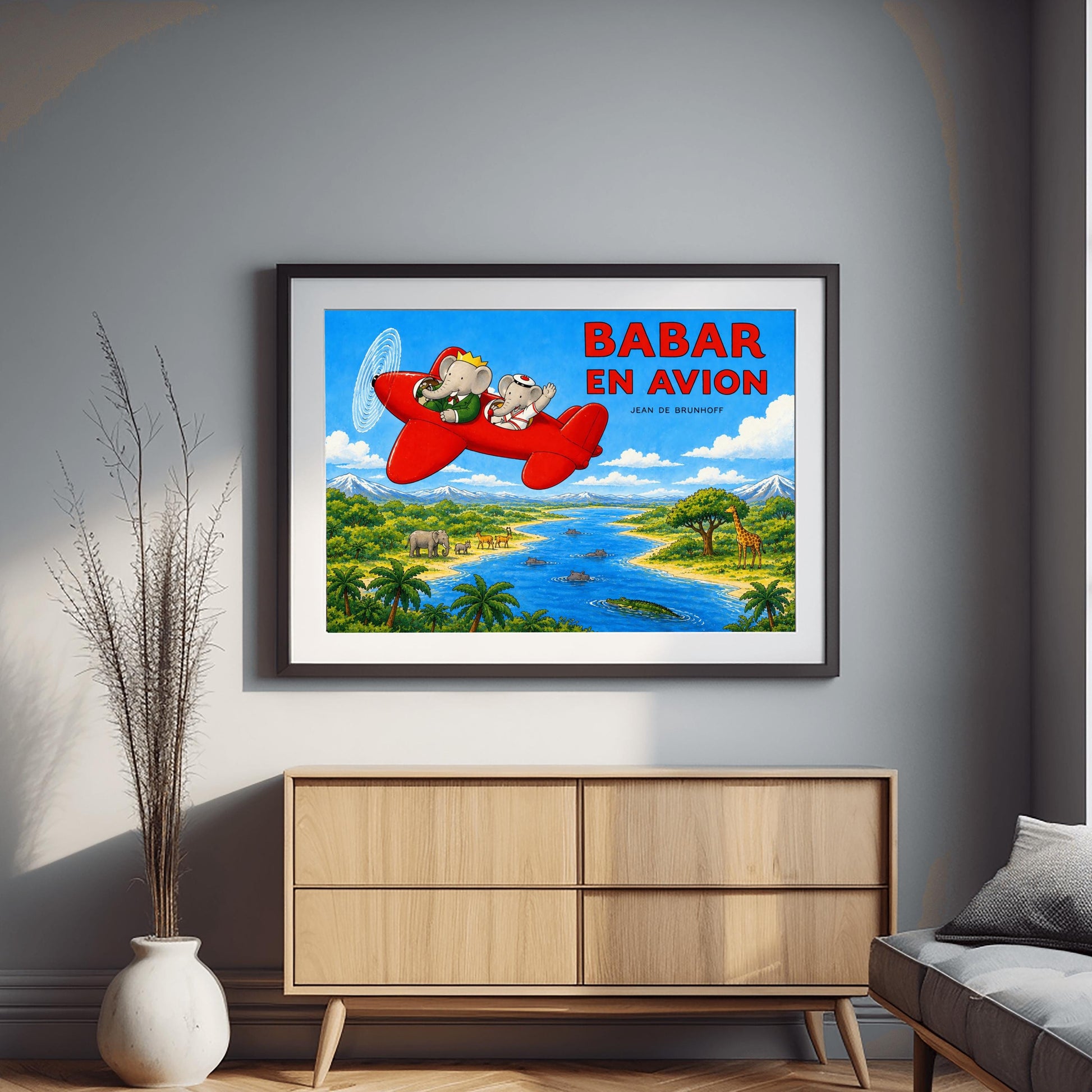 A horizontal watercolor-style illustration of Babar the Elephant flying a bright red propeller plane. Babar wears a green suit and crown, while a smaller elephant in a white sailor suit waves from the back seat. They fly over a vibrant safari landscape with a winding blue river filled with hippos and a crocodile. On the riverbanks are giraffes, antelopes, elephants, and palm trees. Snow-capped mountains rise in the distance under a blue sky. Large red text reads "BABAR EN AVION."