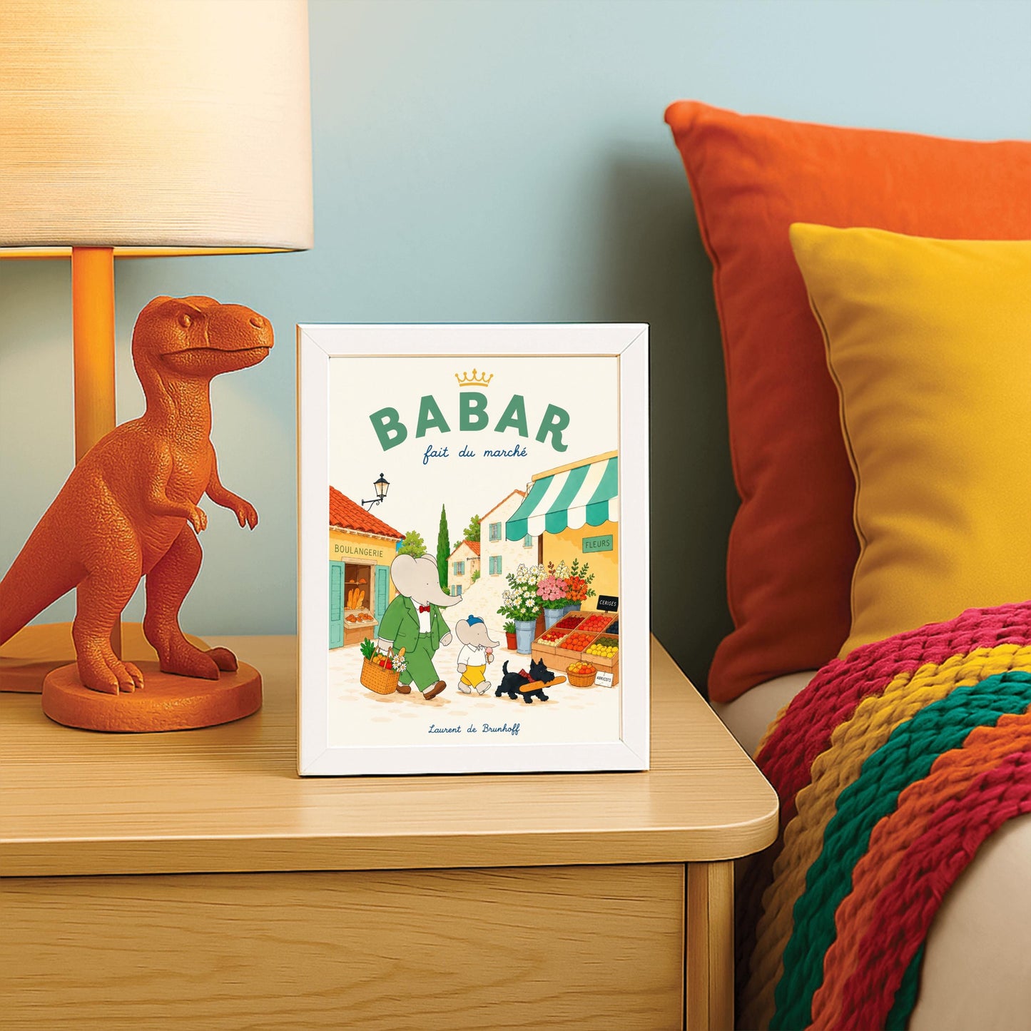 A vertical storybook-style art print of Babar the Elephant walking through a French village street. Babar, in a green suit, carries a basket of groceries. A smaller elephant walks beside him eating a pink ice cream cone, followed by a black dog carrying a baguette. The background features a boulangerie with a yellow facade, a flower stall with pink and white roses, and terracotta-roofed houses under a cream sky. The top reads "BABAR fait du marché" with a gold crown.