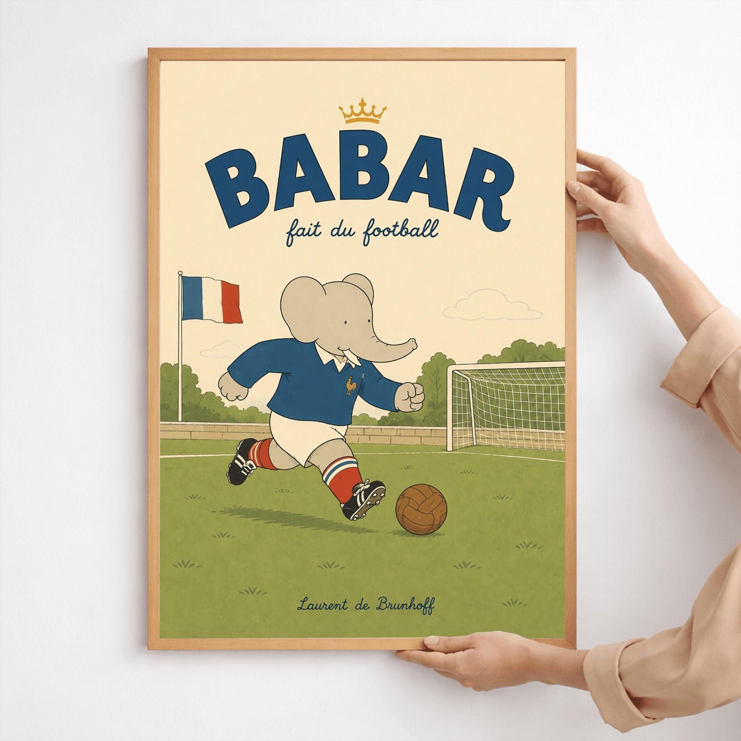 A vertical storybook-style art print featuring Babar the Elephant playing soccer on a green field. Babar wears a blue jersey with a small gold rooster logo, white shorts, red striped socks, and black cleats. He is dribbling a brown leather soccer ball toward a white goal. A French flag flies in the background. The text at the top says "BABAR fait du football" with a small gold crown.