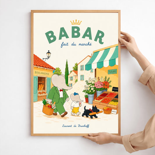 A vertical storybook-style art print of Babar the Elephant walking through a French village street. Babar, in a green suit, carries a basket of groceries. A smaller elephant walks beside him eating a pink ice cream cone, followed by a black dog carrying a baguette. The background features a boulangerie with a yellow facade, a flower stall with pink and white roses, and terracotta-roofed houses under a cream sky. The top reads "BABAR fait du marché" with a gold crown.