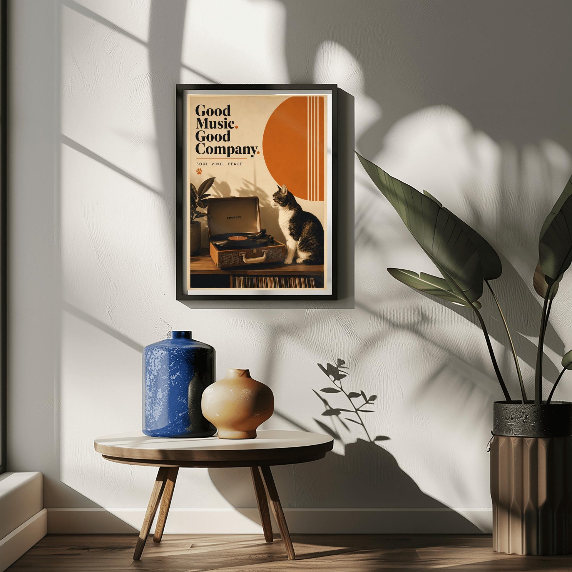 A vertical retro fine art poster titled "Good Music. Good Company." on a wide cream border. The design features a bold black serif heading on the left above the text "SOUL. VINYL. PEACE." and a small orange paw print. In the central artwork, a black and white tuxedo cat sits contentedly next to an open tan Crosley record player spinning a vinyl record. A large orange sun shape with white vertical stripes and a potted rubber plant decorate the background.
