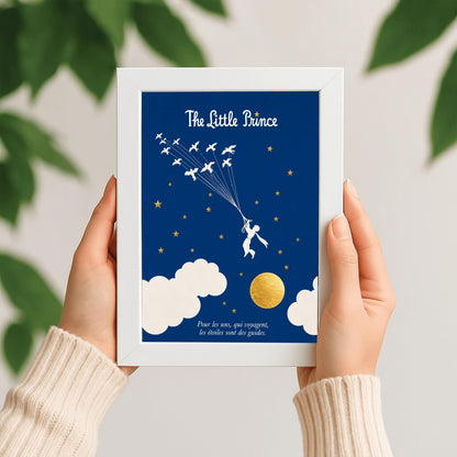 A vertical art print with a deep indigo blue background. A small white silhouette of a boy, the Little Prince, is being pulled upward by a flock of white birds on long strings. The sky is filled with small gold stars and large white clouds at the bottom, next to a textured gold moon. The title "The Little Prince" is at the top in white script, and a French quote is at the bottom in white italics.