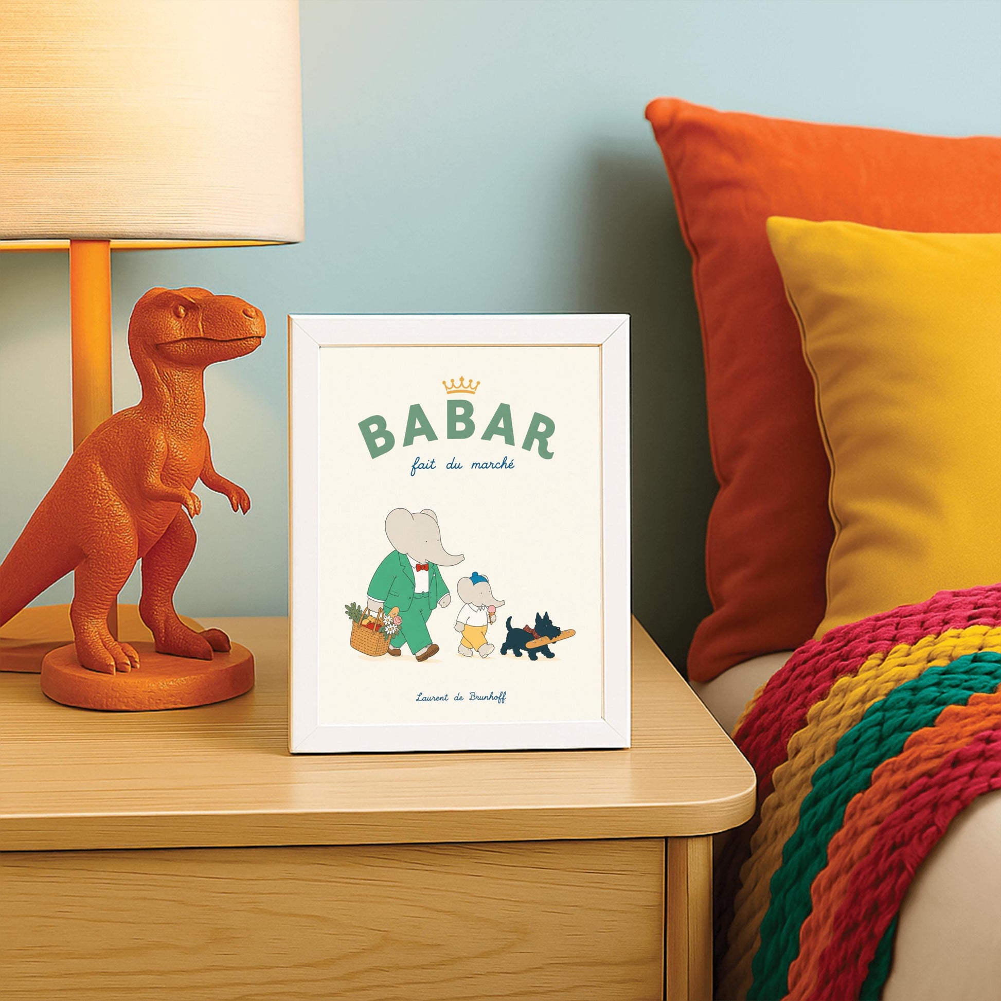 A vertical storybook-style art print of Babar the Elephant walking home from a market against a cream background. Babar is in a green suit and red bow tie, carrying a basket of fruit and flowers. Beside him is a smaller elephant eating an ice cream cone and a small black dog in a plaid collar carrying a baguette. The top text reads "BABAR fait du marché" with a gold crown.
