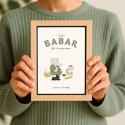 A vertical storybook-style art print of Babar the Elephant having a picnic against a cream background. Babar, in a green suit, sits on a white blanket with a smaller elephant in a blue hat and yellow shorts. A small black dog lies in the foreground. The spread includes a baguette, a bowl of cherries, and a bottle. The top reads "BABAR fait du pique-nique" with a gold crown, and the bottom says "Laurent de Brunhoff."