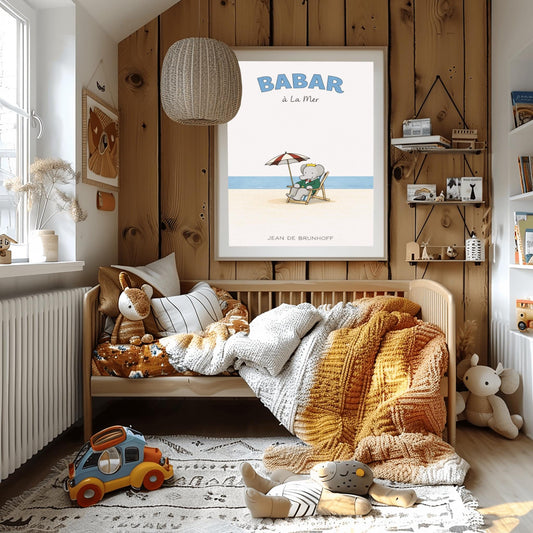 A vertical children's illustration poster titled "BABAR à La Mer" in light blue. In the center, an elephant (Babar) in a green suit and crown is sitting in a wooden lounge chair on a sandy beach. A red and white striped umbrella is behind him. In the background, a thin blue line represents the sea under a white sky. The name "JEAN DE BRUNHOFF" is at the bottom.