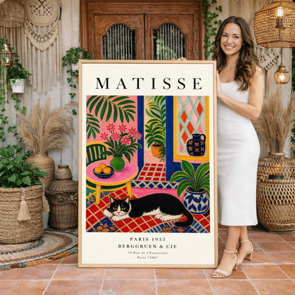 A vertical maximalist art print styled as a Henri Matisse exhibition poster. A black and white tuxedo cat lies across an intricate red, orange, and blue patterned rug. The room features textured bubblegum pink walls and a cobalt blue door frame. To the left, a pink table holds a bowl of oranges and a green vase with pink flowers. To the right, a blue and white floral pot holds a green plant. A diamond-patterned window is visible in the background.