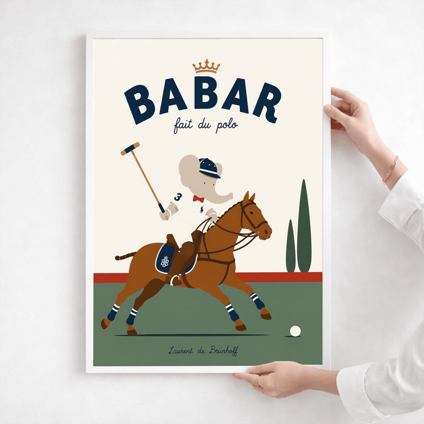 A vertical storybook-style art print of Babar the Elephant playing polo. Babar is riding a chestnut brown horse, wearing a white jersey with the number 3, a red bow tie, and a navy riding helmet. He holds a polo mallet in one hand. The horse has navy and white striped leg wraps. The background features a green field, a red horizon line, and two green cypress trees under a cream sky. The top reads "BABAR fait du polo" with a gold crown.