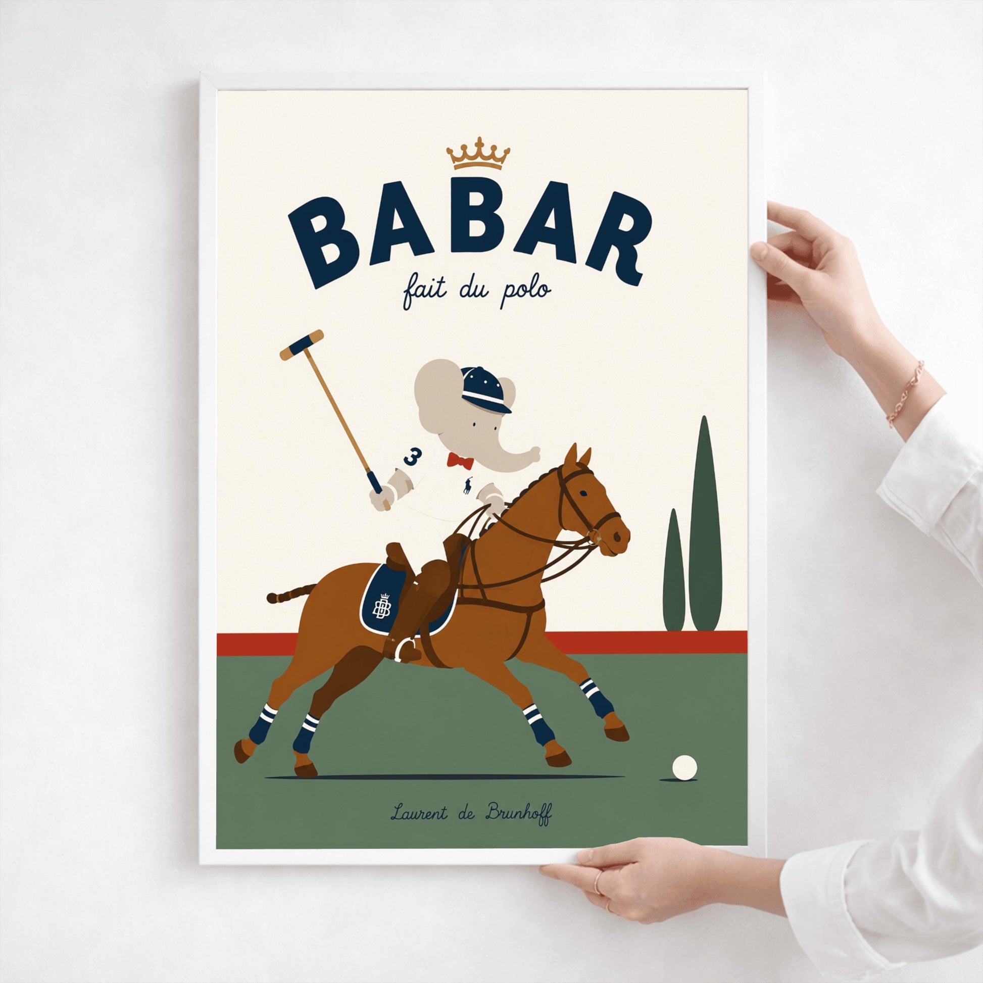 A vertical storybook-style art print of Babar the Elephant playing polo. Babar is riding a chestnut brown horse, wearing a white jersey with the number 3, a red bow tie, and a navy riding helmet. He holds a polo mallet in one hand. The horse has navy and white striped leg wraps. The background features a green field, a red horizon line, and two green cypress trees under a cream sky. The top reads "BABAR fait du polo" with a gold crown.