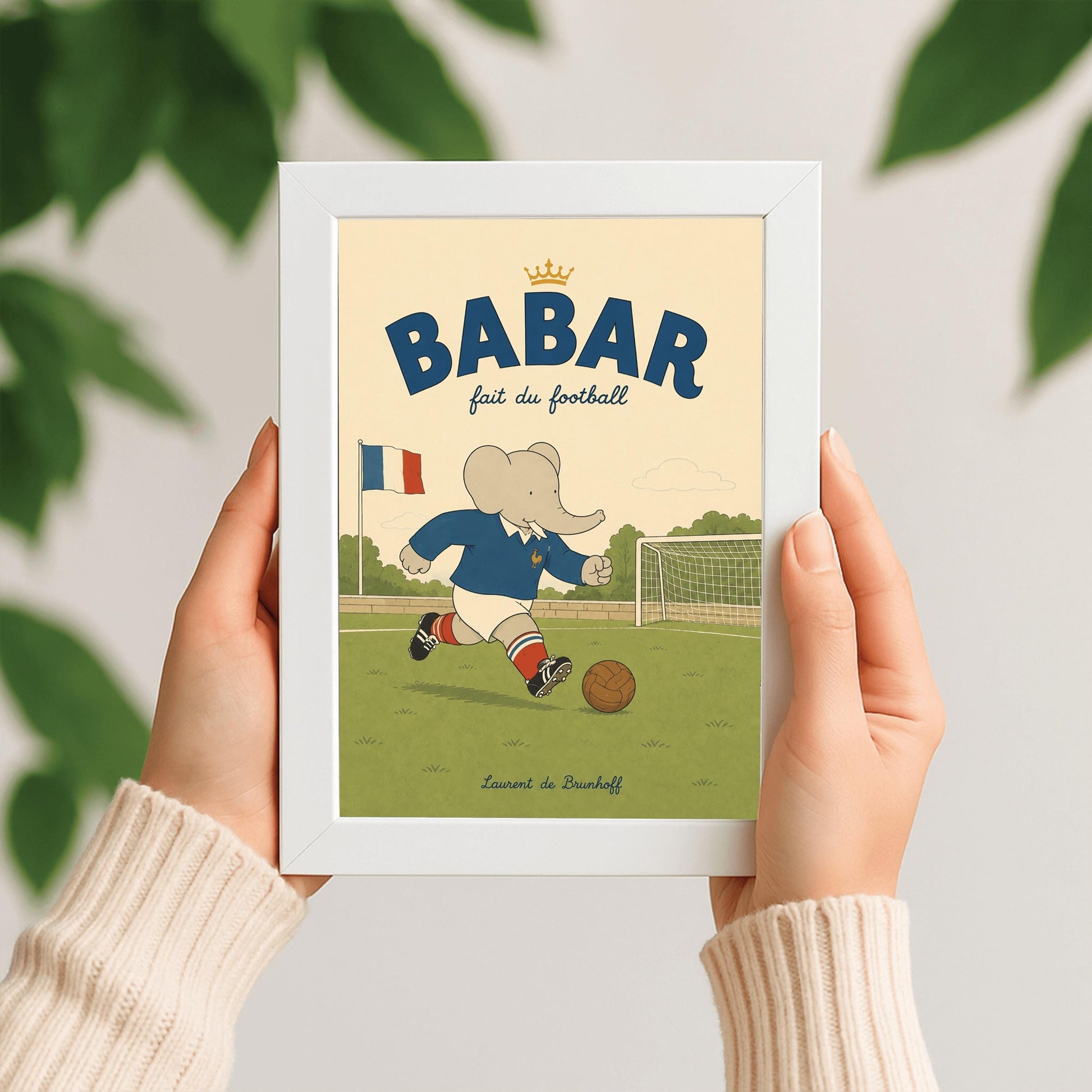 A vertical storybook-style art print featuring Babar the Elephant playing soccer on a green field. Babar wears a blue jersey with a small gold rooster logo, white shorts, red striped socks, and black cleats. He is dribbling a brown leather soccer ball toward a white goal. A French flag flies in the background. The text at the top says "BABAR fait du football" with a small gold crown.
