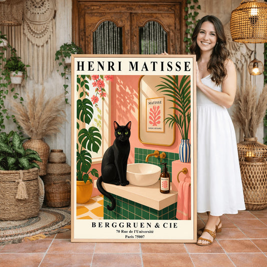 A vertical art print styled as a Henri Matisse exhibition poster. A sleek black cat with bright green eyes sits on a cream-colored bathroom vanity next to a white vessel sink. The bathroom features blush pink walls and emerald green tiles. A gold-framed mirror reflects a Matisse leaf print. Lush green plants and pink flowers frame the scene, and dappled sunlight creates shadows on the wall. The poster has bold black text at the top and bottom with the gallery name Berggruen & Cie.