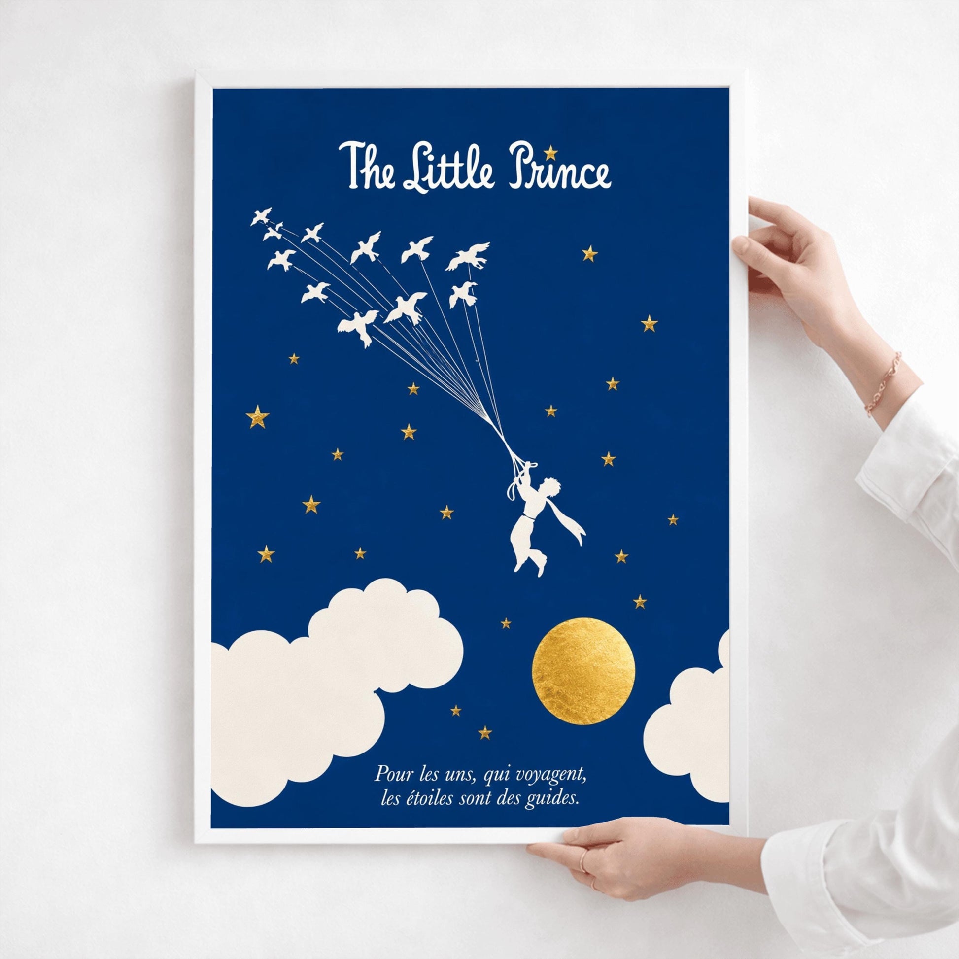 A vertical art print with a deep indigo blue background. A small white silhouette of a boy, the Little Prince, is being pulled upward by a flock of white birds on long strings. The sky is filled with small gold stars and large white clouds at the bottom, next to a textured gold moon. The title "The Little Prince" is at the top in white script, and a French quote is at the bottom in white italics.