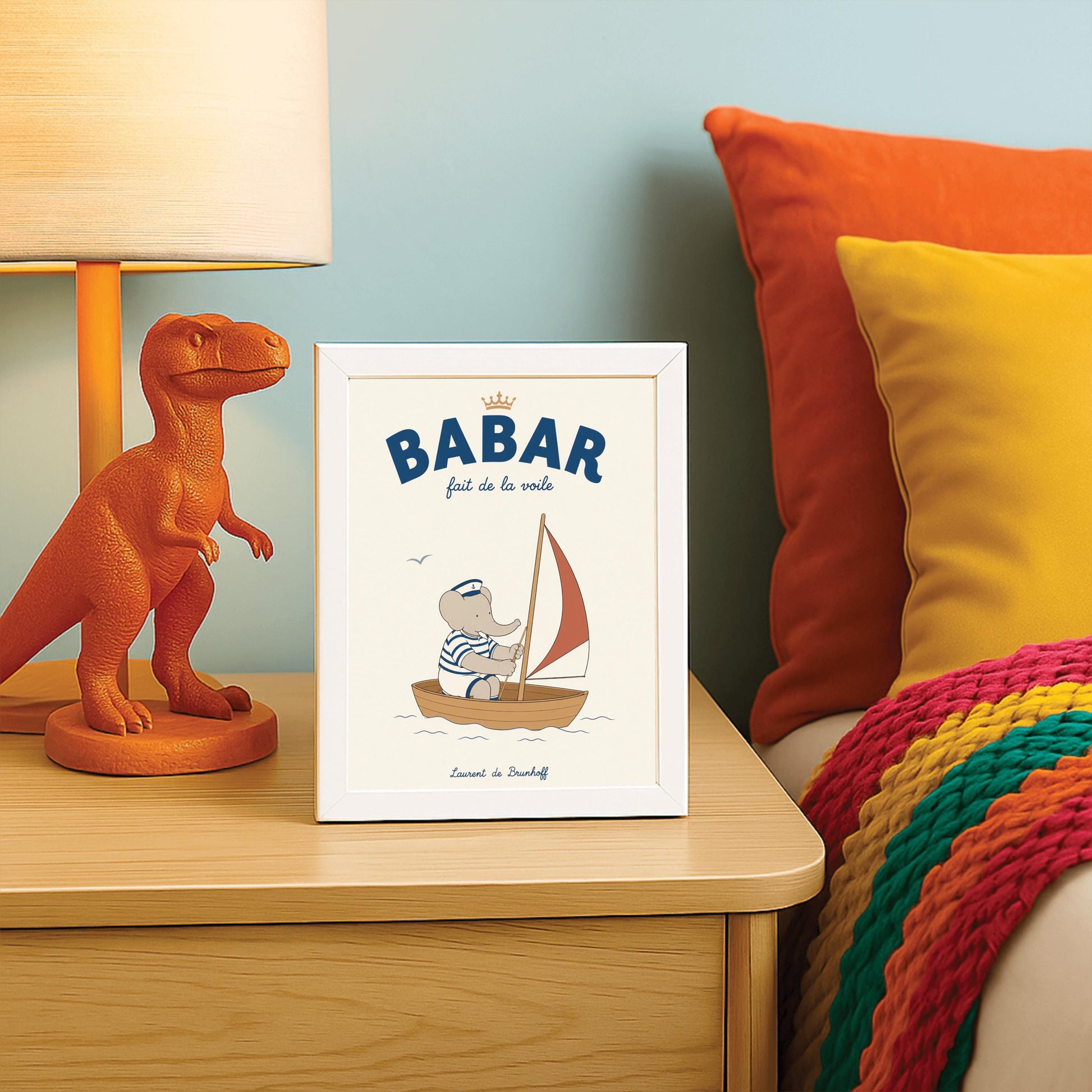 A vertical storybook-style art print featuring Babar the Elephant sailing a small boat. Babar wears a blue and white striped shirt and a white sailor's hat with an anchor. He is sitting in a brown wooden boat with a red and white sail. A single seagull flies in the creamy off-white sky. The top reads "BABAR fait de la voile" with a small gold crown.