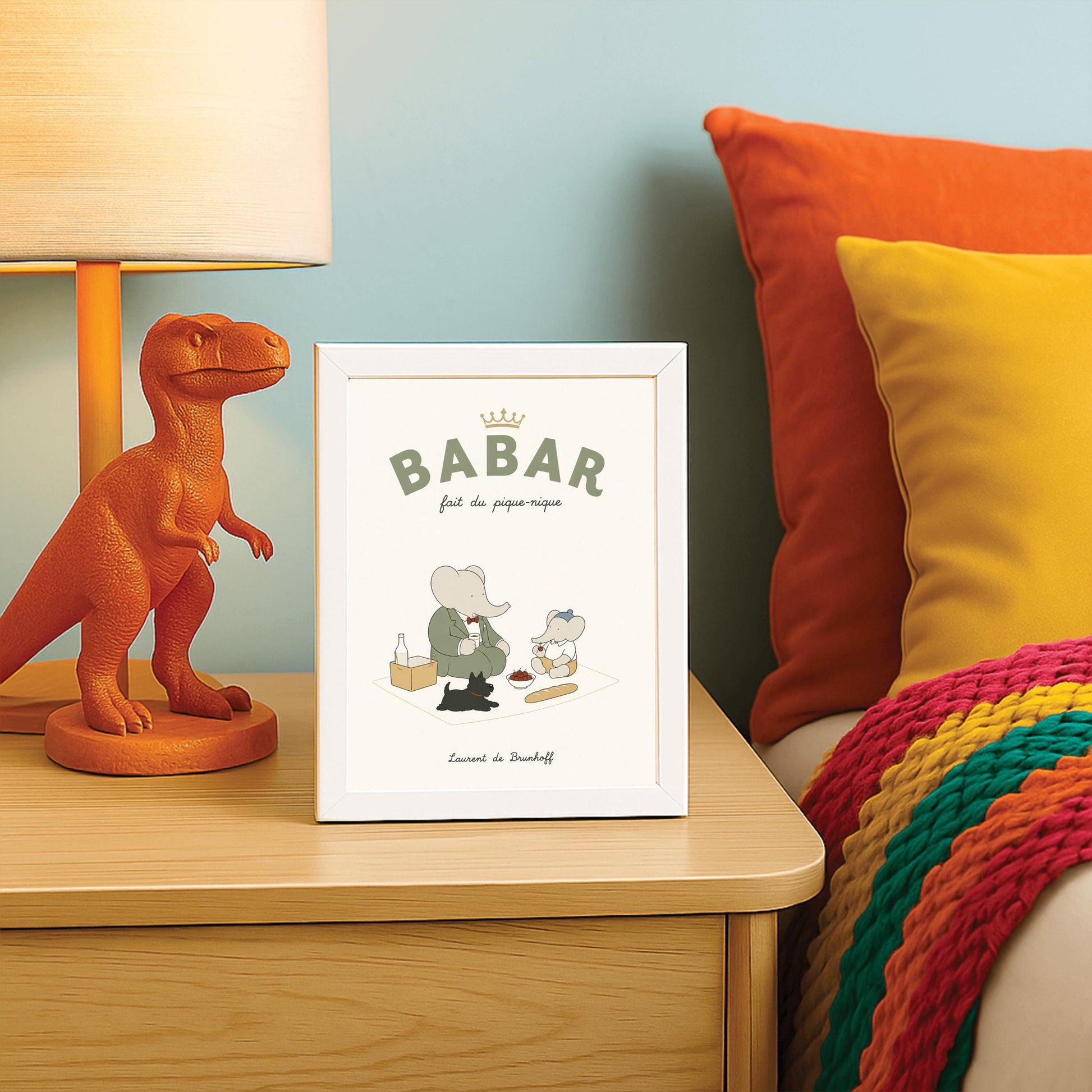A vertical storybook-style art print of Babar the Elephant having a picnic against a cream background. Babar, in a green suit, sits on a white blanket with a smaller elephant in a blue hat and yellow shorts. A small black dog lies in the foreground. The spread includes a baguette, a bowl of cherries, and a bottle. The top reads "BABAR fait du pique-nique" with a gold crown, and the bottom says "Laurent de Brunhoff."