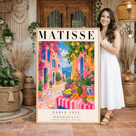 A vertical maximalist art print styled as a Henri Matisse exhibition poster. The scene depicts a vibrant, colorful street in a Mediterranean coastal town. In the foreground, a table with a pink and white striped cloth is piled high with lemons, oranges, and a pineapple. Hot pink and yellow buildings with blue shutters line a cobblestone path that leads toward a blue sea under a bright sky. Lush green trees with yellow lemons and pink flowers frame the view.