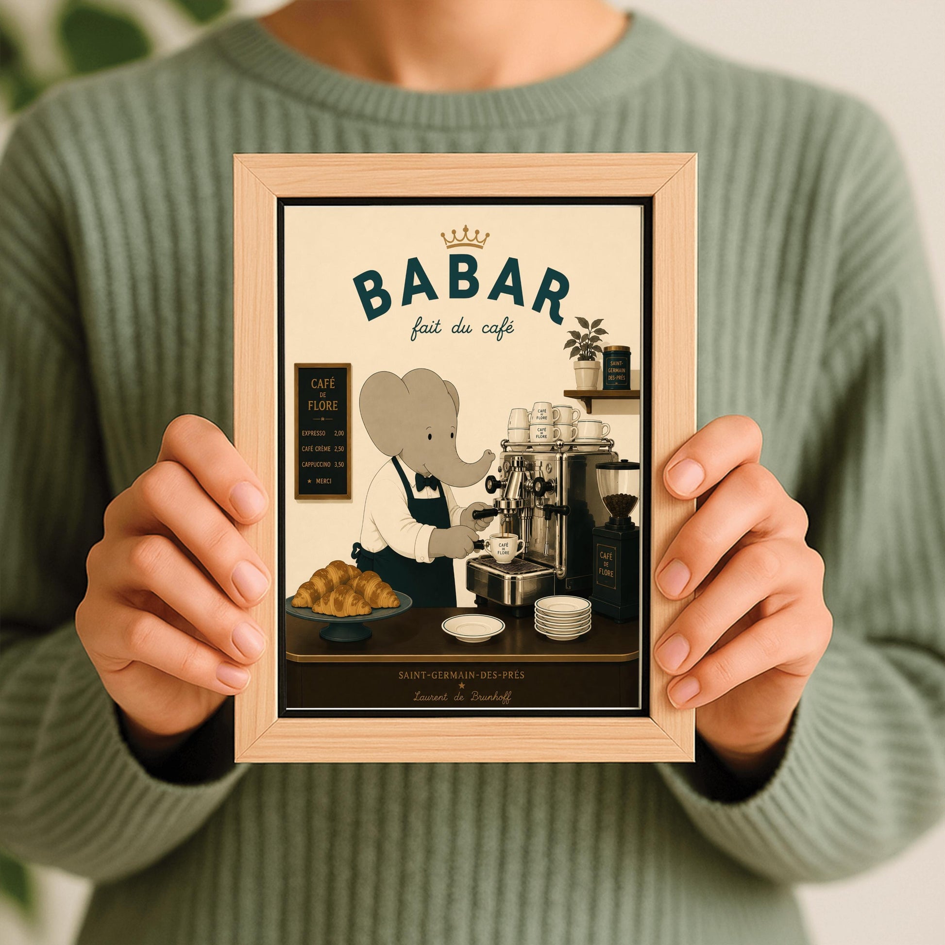 A vintage-style illustration of Babar the Elephant wearing a white shirt, black bowtie, and dark apron, working as a barista. He is operating a classic silver espresso machine in a Parisian café. The scene includes a plate of croissants, stacks of "Café de Flore" cups, and a menu board. The text at the top reads "BABAR fait du café" with a gold crown, and the bottom features "Saint-Germain-des-Prés" and the name "Laurent de Brunhoff."