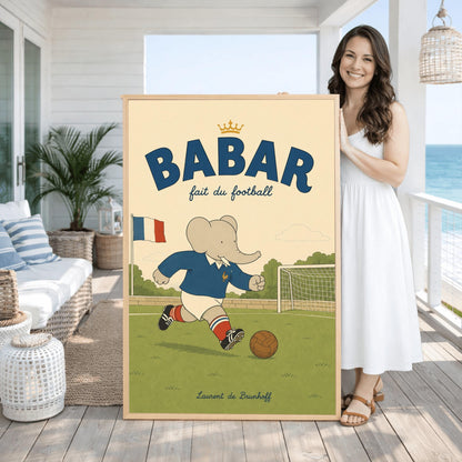 A vertical storybook-style art print featuring Babar the Elephant playing soccer on a green field. Babar wears a blue jersey with a small gold rooster logo, white shorts, red striped socks, and black cleats. He is dribbling a brown leather soccer ball toward a white goal. A French flag flies in the background. The text at the top says "BABAR fait du football" with a small gold crown.