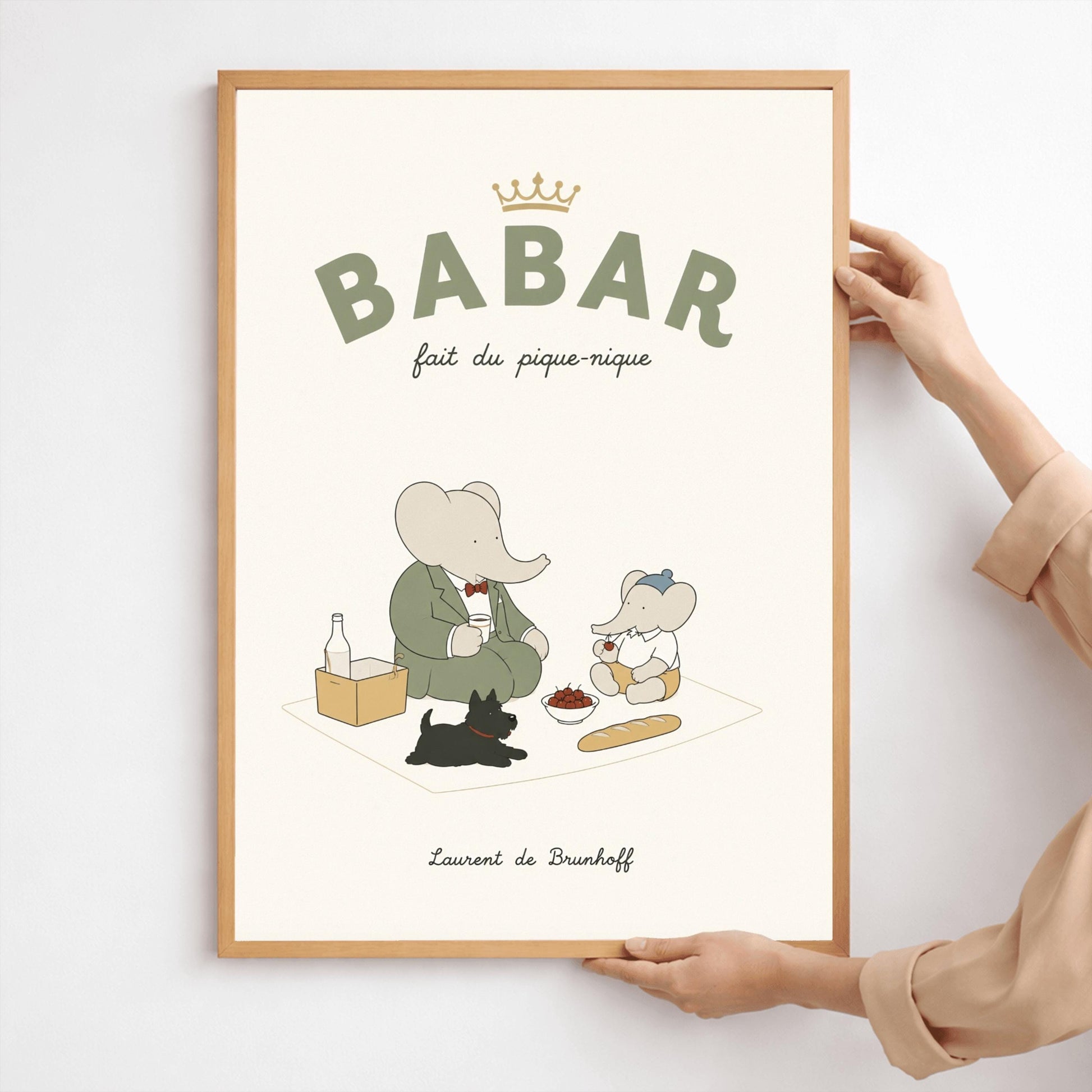 A vertical storybook-style art print of Babar the Elephant having a picnic against a cream background. Babar, in a green suit, sits on a white blanket with a smaller elephant in a blue hat and yellow shorts. A small black dog lies in the foreground. The spread includes a baguette, a bowl of cherries, and a bottle. The top reads "BABAR fait du pique-nique" with a gold crown, and the bottom says "Laurent de Brunhoff."
