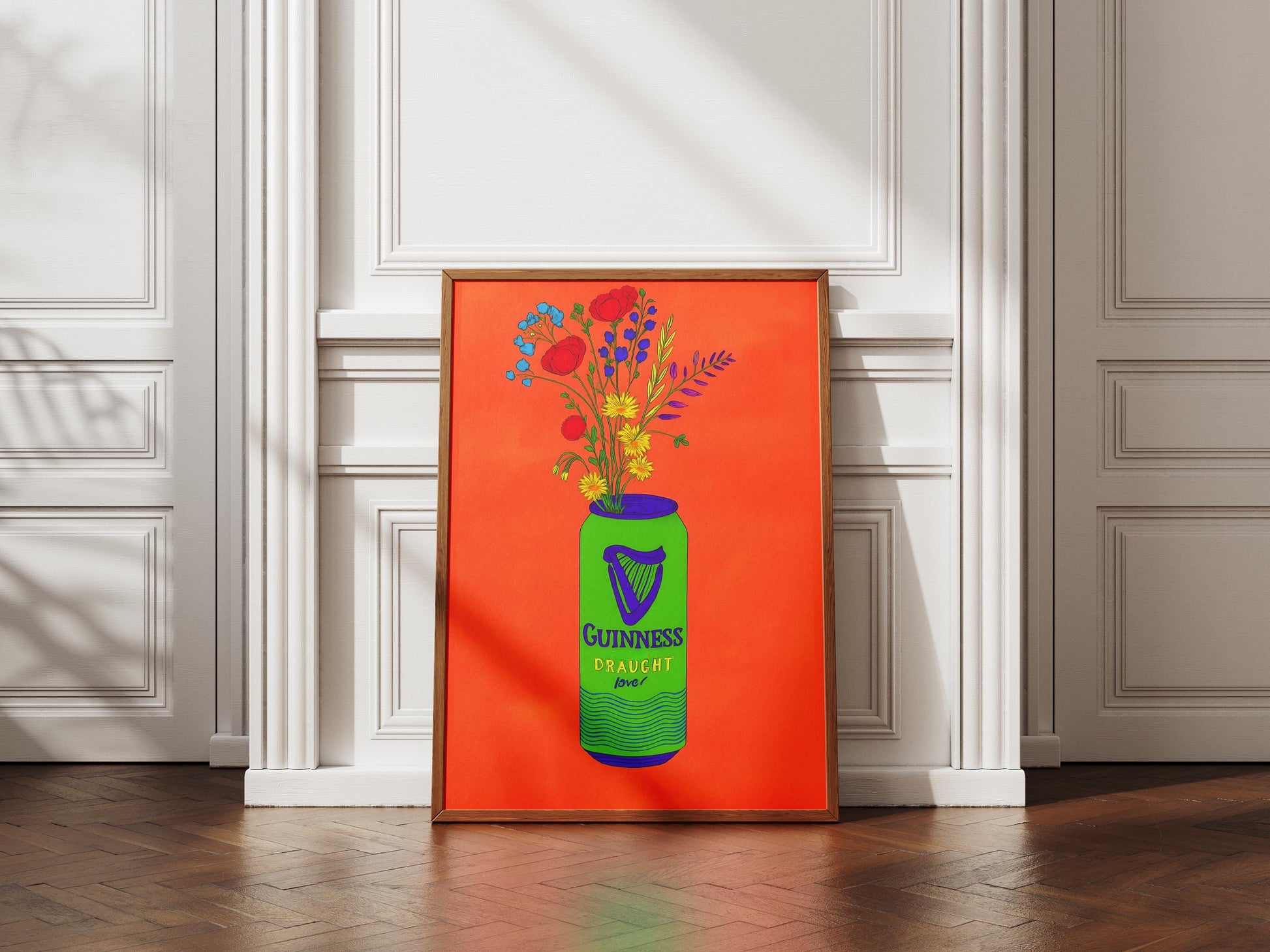 Framed artwork of a green Guinness bottle with flowers against an orange background, displayed in a room with white walls and wooden floor.