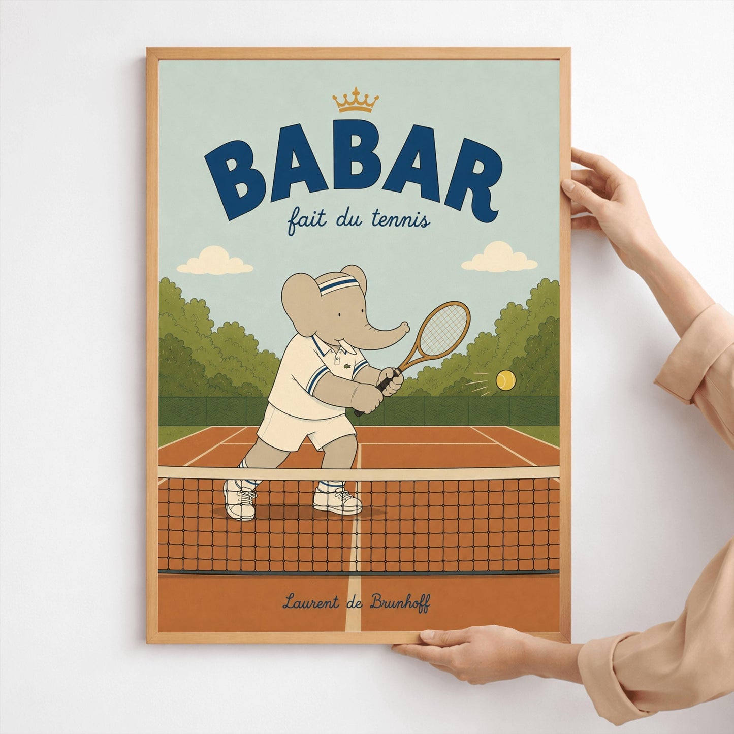 A vertical storybook-style art print featuring Babar the Elephant playing tennis. Babar is seen behind a tennis net on an orange clay court, holding a wooden racket in a backhand swing. He wears a white polo with blue stripes, white shorts, and a blue-and-white striped headband. A yellow tennis ball is in mid-air. The background has green trees and a light blue sky. The top reads "BABAR fait du tennis" with a small gold crown.