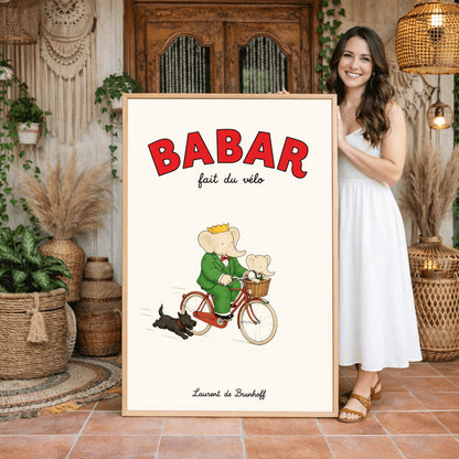 A vertical retro fine art exhibition poster titled "BABAR fait du vélo" on a wide cream border. The top "BABAR" text is printed in a bold red font. The illustration shows an elephant wearing a crown and green suit riding a vintage red bicycle. A smaller baby elephant is sitting inside a brown wicker basket on the handlebars, and a small black dog runs along beside the bike. The name "Laurent de Brunhoff" anchors the bottom.
