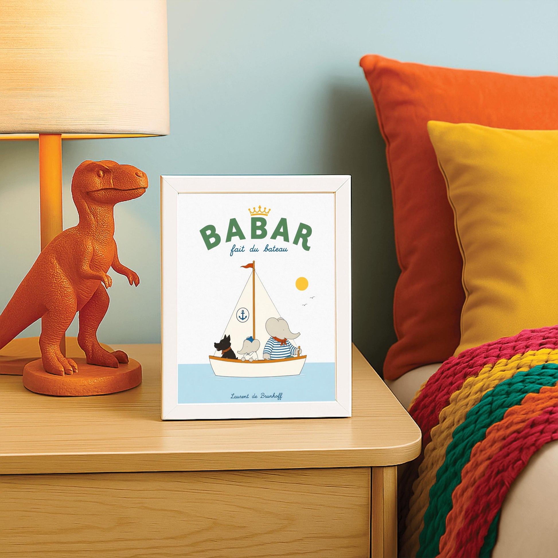 A vertical storybook-style art print of Babar the Elephant sailing a boat against a white background. Babar, wearing a blue and white striped shirt and a red neckerchief, sits in a white boat with a smaller elephant and a black dog. The sailboat has a white sail with a blue anchor logo and a red flag on top. A yellow sun and two small birds are in the sky. The top reads "BABAR fait du bateau" with a gold crown.