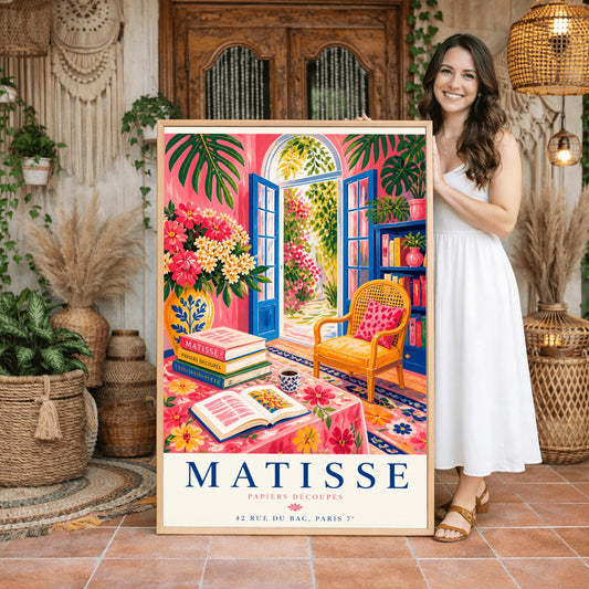A vertical maximalist art print styled as a Henri Matisse exhibition poster. The scene shows a vibrant interior reading room with hot pink walls and cobalt blue French doors open to a sunlit garden. A yellow cane chair sits next to a bookshelf. In the foreground, a table covered in a pink floral cloth holds a stack of art books, an open book, and a blue coffee mug. Large monstera leaves and various flowers frame the room. Bold black and red text at the bottom reads "MATISSE PAPIERS DÉCOUPÉS"